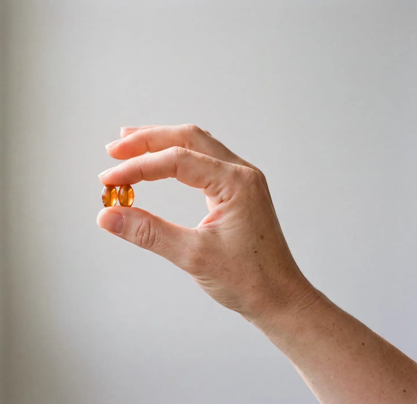 A hand holding two amber softgel capsules against a plain pale wall in soft daylight