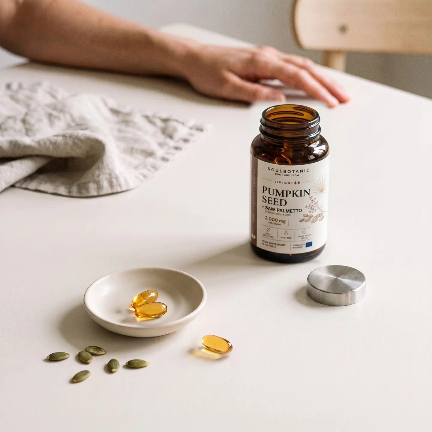 Une photo du pot de Soulbotanic « Graines de citrouille + palmier nain » ouvert sur une table de cuisine claire, baignée d’une douce lumière du jour, son bouchon en acier brossé posé à côté, et au premier plan, un petit plat en céramique contenant des gélules dorées parsemées de graines de citrouille, avec une main posée sur la table, floue à l’arrière-plan.